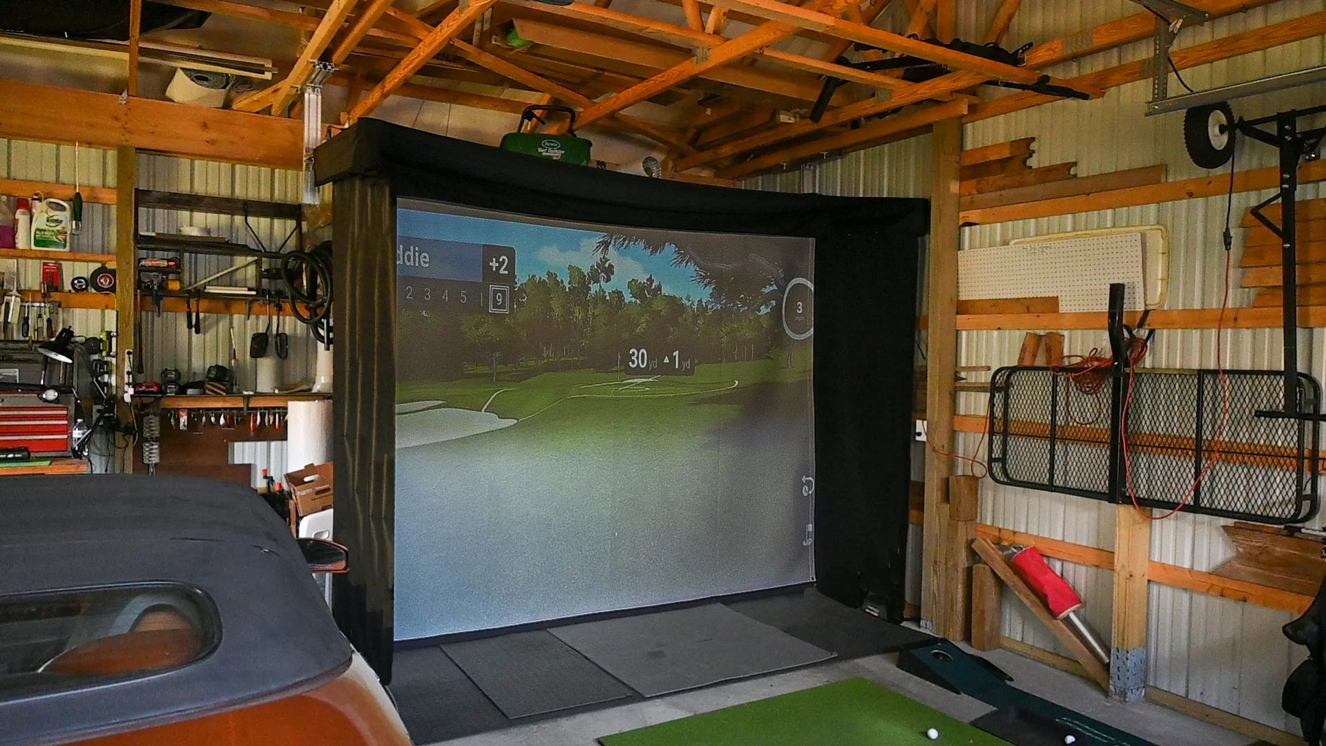 Indoor golf simulator setup in a garage with a screen displaying a golf course. SportScreen Vanish Lite Retractable Golf Simulator enclosure.